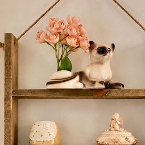 May include: A three-tiered wooden wall shelf suspended by a jute rope. The top shelf holds a ceramic Siamese cat figurine with pink flowers. The middle shelf features a small patterned bowl and a lidded trinket box. The bottom shelf displays a framed portrait and a small floral vase.