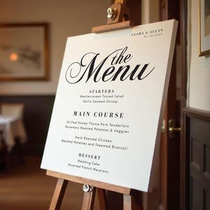 May include: A white sign with black text on an easel, announcing the wedding menu for Clara & Allan on July 18, 2024. The menu includes starters, main course, and dessert options.