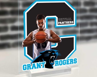 Op de afbeelding: Een transparante acrylplaat met een zwarte en blauwe afbeelding van een basketballer. De speler houdt een basketbal vast en draagt een shirt met nummer 15. De plaat heeft de tekst "CENTRAL PANTHERS" en "GRANT ROGERS" in blauw.
