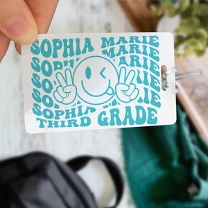 May include: White luggage tag with a turquoise smiley face with peace signs. The tag has the text "Sophia Marie Third Grade" repeated in a turquoise swirl pattern.