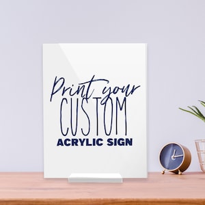 May include: A white acrylic sign with the text "Print your CUSTOM ACRYLIC SIGN" in navy blue script. The sign is displayed on a wooden surface with a small clock and decorative gold wire basket.