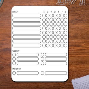 May include: A white habit tracker with rounded corners, featuring sections for daily, weekly, and monthly goals. It includes spaces for checkmarks and lines for writing, with the days of the week labeled across the top. The tracker is on a wooden surface.