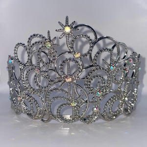 May include: A silver crown with an elaborate openwork design. The crown features swirling patterns, star-shaped accents, and iridescent gemstones. This tiara is suitable for special occasions and events.