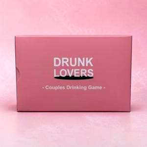 May include: A rectangular pink box for a drinking game. The front features the white text DRUNK LOVERS in a bold sans-serif font, with a black shadow effect underneath the word LOVERS, and the text - Couples Drinking Game - below it.