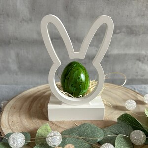 Könnte beinhalten: Weißer hölzerner Osterhasen-Eierhalter mit einem grünen marmorierten Osterei darin. Der Halter steht auf einer Holzscheibe mit grünen Blättern und weißen Lichtern.
