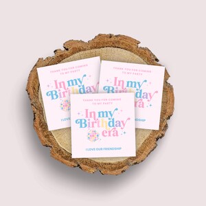 May include: Three pink thank you cards with the text "In my Birthday era" in colourful letters and a disco ball graphic. The text "Thank you for coming to my party" is also on the cards. The cards are on a wooden slice.