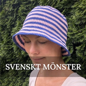 以下が含まれることがあります： 青とピンクのストライプのクロシェのつば広帽子。帽子は白いシャツを着た人がかぶっています。画像には「SVENSKT MÖNSTER」というテキストが表示されています。