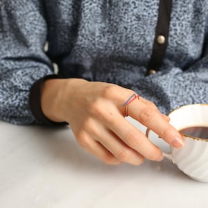 Könnte beinhalten: Ein goldener Ring mit einem roten und blauen Emaille-Design. Der Ring wird am Zeigefinger einer Hand getragen, die eine Tasse Kaffee hält.