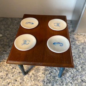 Puede incluir: Cuatro platos de cerámica blanca con diseños de cangrejos azules y blancos están dispuestos sobre una mesa de madera con patas pintadas de azul.