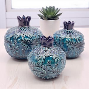 May include: Three ceramic pomegranate sculptures in varying sizes, featuring a teal and blue glaze with intricate floral and leaf patterns. Each piece has a dark blue crown-like top. A small succulent in a white pot is in the background.
