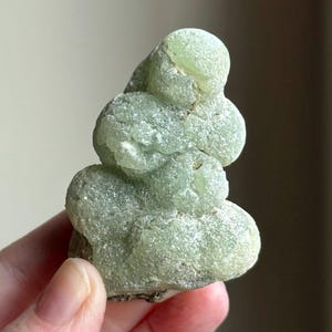 May include: A light green prehnite crystal cluster. The mineral has a bubbly, rounded form with a textured surface. The color is a pale, translucent green, with some areas appearing more opaque. The crystal is held in a hand.