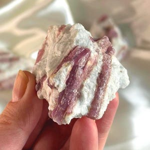 May include: A hand holds a rough, natural stone. The stone is a mix of white and pink, with the pink appearing in long, crystalline formations throughout the white matrix. The overall texture is coarse and uneven.