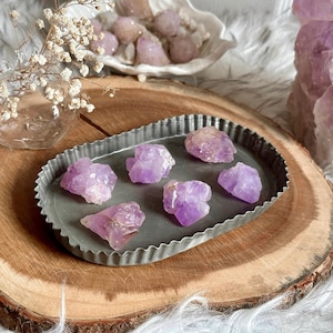 May include: Six small, purple amethyst crystals on a gray metal tray. The tray is on a wooden surface.