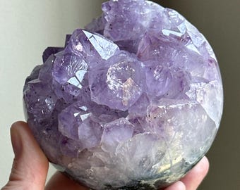 May include: A spherical amethyst geode with a polished exterior and a crystalline interior. The geode features a range of purple hues, from light lavender to deep violet, with sparkling crystal formations. The base of the geode has a dark, natural appearance.