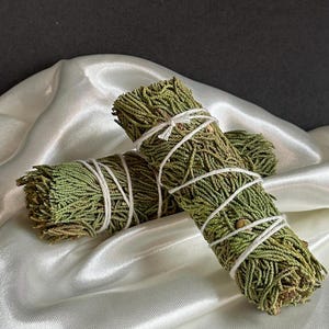May include: Two bundles of dried green herbs, tightly wrapped with white twine. The bundles are resting on a white, silky fabric, with a dark background. The herbs have a natural, textured appearance.