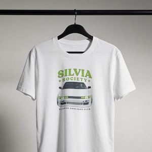 May include: A white short-sleeve t-shirt featuring a graphic of a white car with lime green headlights. Above the car, the text reads SILVIA SOCIETY in lime green. Below the car, the text reads RUBBER HOOLIGAN CLUB in black capital letters.