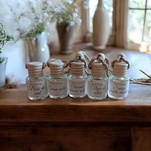 Peut inclure: Cinq petits flacons en verre avec des bouchons en bois et des cordes sont disposés sur une surface en bois. Chaque flacon porte une étiquette avec un nom de parfum différent : Bois de oud, Cerise noire, Caramel beurre sale, Fleur de coton, et Sauge basilic ment.