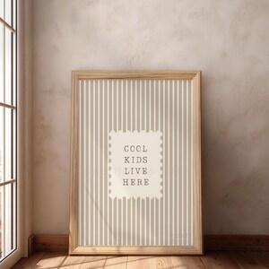 May include: A framed print with a light wood frame. The print has a striped background in shades of beige and white. A white rectangle in the center features the text "COOL KIDS LIVE HERE" in a simple font. The print is leaning against a wall.