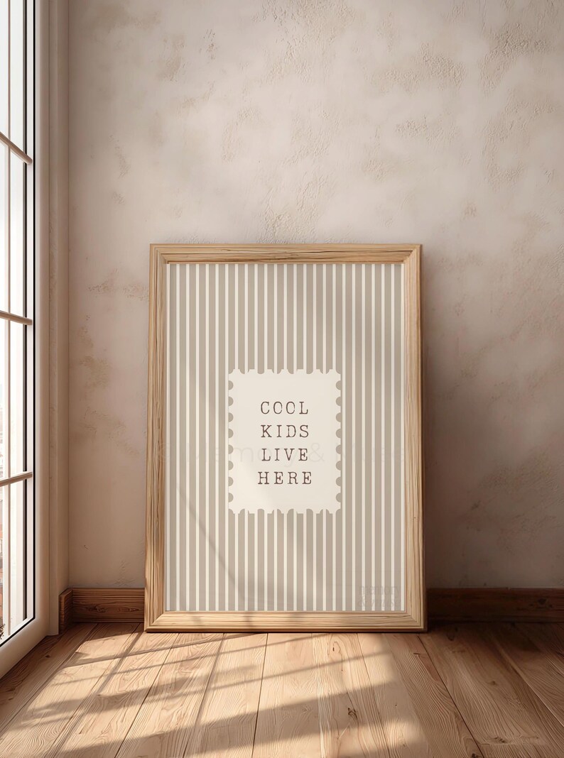 May include: A framed print with a light wood frame. The print has a striped background in shades of beige and white. A white rectangle in the center features the text "COOL KIDS LIVE HERE" in a simple font. The print is leaning against a wall.