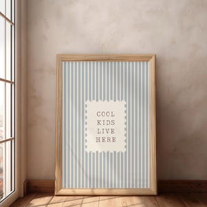 May include: A framed print with a light blue and white striped background. The print features the text "COOL KIDS LIVE HERE" in a stamp-like design. The frame is made of light-colored wood, and the print is leaning against a wall.