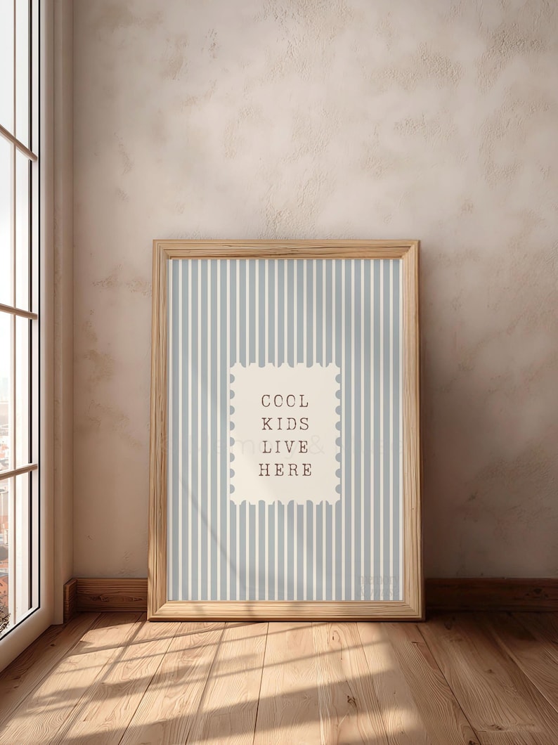 May include: A framed print with a light blue and white striped background. The print features the text "COOL KIDS LIVE HERE" in a stamp-like design. The frame is made of light-colored wood, and the print is leaning against a wall.