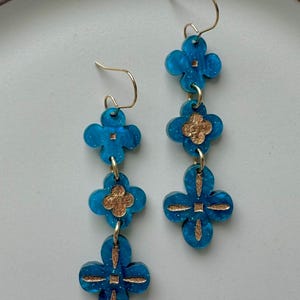 May include: A pair of dangling earrings featuring a three-tiered clover design. The earrings are a vibrant turquoise blue with gold-colored accents and gold-tone ear wires. The clover shapes are connected by small gold rings.