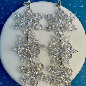 Puede incluir: Dos pares de pendientes de plata con forma de copo de nieve y detalles de purpurina iridiscente. Cada pendiente presenta tres formas de copo de nieve unidas por pequeños aros plateados. Se muestran sobre una superficie circular blanca sobre fondo azul estrellado.