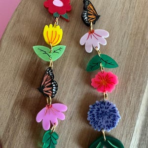 May include: A pair of long, colorful earrings featuring a variety of flowers, leaves, and butterflies. The earrings are made of acrylic and have a gold-tone finish.