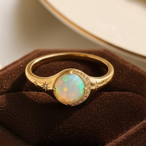May include: A gold ring featuring a round, iridescent opal stone set in the center. The band is decorated with small engraved star motifs and a crescent moon shape surrounding the stone, creating a celestial design on the polished metal surface.