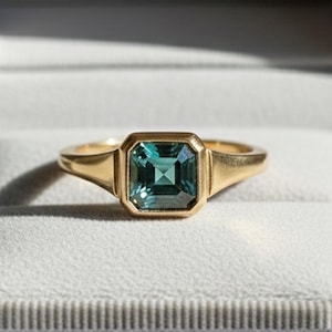 Peut inclure: Une bague en or avec une pierre précieuse carrée de couleur teal sertie dans un sertissage à lunette. La bague est présentée dans un écrin gris ouvert avec un intérieur de couleur claire. La bande de la bague a un design simple et classique.