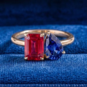 Könnte beinhalten: Ein Goldring mit einem rechteckigen, im Smaragdschliff geschliffenen roten Edelstein und einem birnenförmigen blauen Edelstein, die nebeneinander auf einer dünnen Goldschiene sitzen, präsentiert in einer tiefblauen Samtschmuckschatulle mit Klappdeckel.