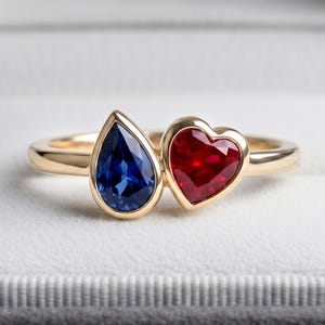 Può includere: Un anello d'oro con una gemma blu a forma di pera e una gemma rossa a forma di cuore. L'anello è presentato in un cofanetto grigio con interno bianco. Le pietre preziose sono il punto focale dell'anello, incastonate in una fascia d'oro.