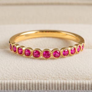Pode incluir: Um anel de banda dourada com uma fila de pedras redondas de cor rosa vibrante, engastadas em biséis individuais com uma textura milgrain nas bordas. O anel é exibido horizontalmente, destacando o engaste circular consistente.