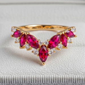 Può includere: Un anello in oro a forma di V con una serie di rubini rosso vivo taglio marquise che si alternano a piccoli diamanti bianchi rotondi e trasparenti. L'anello è montato su una fascia in oro giallo lucido ed è esposto in un cofanetto in velluto grigio.