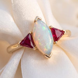 May include: A gold ring featuring a central marquise-cut opal with iridescent flashes of color, flanked by two triangular-cut deep red rubies. The stones are set in a polished gold band, creating a symmetrical design with a vibrant gemstone display.