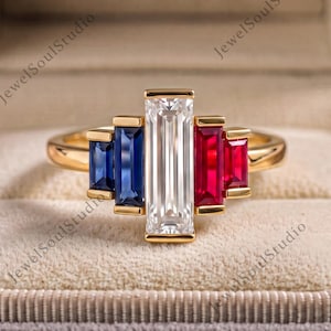 May include: A gold ring featuring a central vertical baguette-cut clear stone flanked by two blue baguette-cut stones on the left and two red baguette-cut stones on the right, all set in a polished gold band with a multi-stone stepped design.