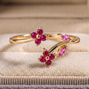 May include: A gold open-style ring featuring two floral motifs composed of round, deep pink rubies and two marquise-cut pink stones. The band is polished gold, and the design showcases a delicate arrangement of gemstone petals and leaves.
