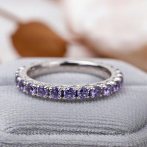 Op de afbeelding: Een zilveren ring met een doorlopende rij kleine, ronde, paarse edelstenen. De ring wordt getoond op een grijze fluwelen ondergrond, met een vage achtergrond in neutrale tinten. De ring is waarschijnlijk een trouw- of verlovingsring.