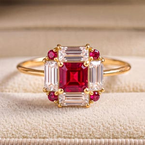 May include: A gold ring featuring a central square-cut red ruby surrounded by four baguette-cut clear diamonds and four small round-cut red rubies. The stones are set in a yellow gold band, creating a geometric cluster design on the ring face.