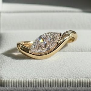 May include: A marquise-cut diamond ring in a gold band, presented in a grey velvet ring box. The diamond is the central feature, catching the light. The gold band curves around the diamond, offering a contemporary design.