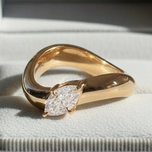 Op de afbeelding: Een gouden ring met een marquise-geslepen diamant, gezet in een uniek, vloeiend ontwerp. De ring wordt gepresenteerd in een open grijze sieradendoos, die de elegante vorm en schittering benadrukt.