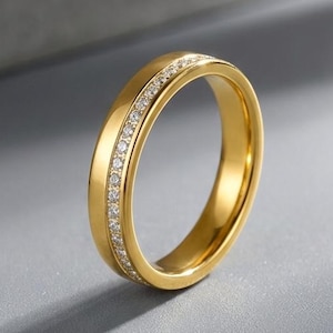 Peut inclure: Une bague dorée ornée d'une rangée de petits diamants transparents. La bague est posée sur un fond gris, avec un design simple et élégant. Il s'agit d'un bijou.