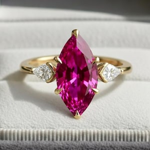 May include: A gold ring with a large, marquise-cut, hot pink gemstone as the centerpiece. Two smaller, triangular, clear gemstones flank the main stone. The ring is displayed in a gray velvet ring box.