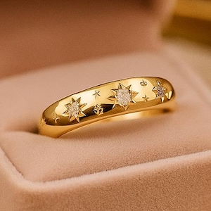 May include: A gold ring with star and diamond detailing sits in a pink velvet ring box. The ring has star-shaped designs and small diamonds. The open ring box displays the ring against a soft, neutral background, creating a luxurious feel.