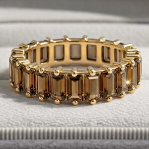 May include: A gold eternity band featuring a continuous row of rectangular, baguette-cut smoky brown gemstones set in a prong setting. The ring is displayed inside a gray velvet-lined jewelry box with a light gray interior cushion.