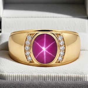 Puede incluir: Anillo de oro con una gran gema de rubí estrella ovalada en el centro. El anillo está flanqueado por pequeños diamantes redondos a cada lado. Presentado en una caja de anillo de terciopelo gris.