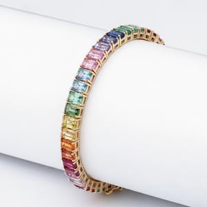 May include: A gold-toned bracelet featuring a row of rectangular, multi-colored gemstones. The gemstones are arranged in a rainbow spectrum, transitioning from red to purple. The bracelet is displayed on a white background.
