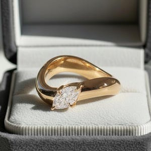 May include: A gold ring featuring a marquise-cut diamond, set in a distinctive, flowing design. The ring is presented in an open, grey velvet ring box, highlighting its elegant form and sparkling gemstone. The ring's design is modern and eye-catching.