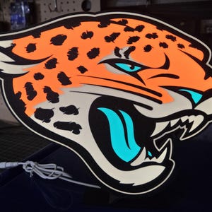 May include: A large, stylized jaguar head logo in orange, black, white, and teal. The jaguar's head is orange with black spots, outlined in black and white. The eyes and tongue are teal. The logo is likely for a sports team.