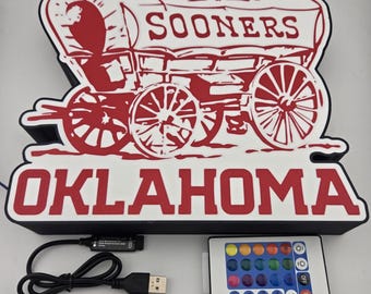 May include: A red and white Oklahoma Sooners sign featuring a covered wagon design. The sign includes the words "SOONERS" and "OKLAHOMA". A remote control and USB cable are included.